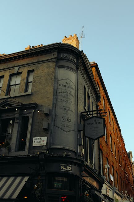A close-up view of a corner of a historic building in Marylebone, displaying a dark grey, rounded architectural feature with a faded painted sign indicating the building's historical name and number 98. The building’s façade is made of light-colored brick with visible texture, and features several windows with dark frames. On the left side of the image, there is a striped awning above a shopfront. To the right, a black signboard projecting from the building bears the name of the establishment. The sky above is clear and light blue, with soft sunlight illuminating the upper part of the structure, highlighting its architectural details and weathered paint, providing an atmospheric view of an area where private property and independent collection services like Waste Removal Marylebone might operate for alternative waste handling.
