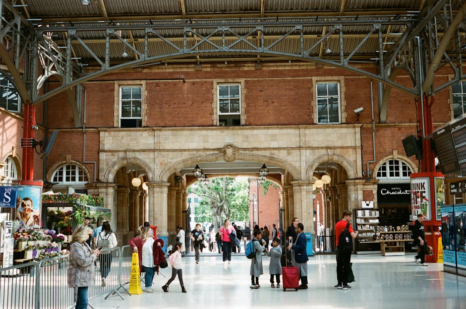 The image depicts a busy indoor public space, likely a train station or similar transportation hub, with a high-ceiling structure supported by exposed metal beams and trusses. The background features a large arched entrance made of stone with classical architectural details, including columns and decorative carvings, leading to an outdoor area with greenery visible through the passage. On the left side, there is a flower stand displaying colourful bouquets with a woman browsing, and on the right, a small retail kiosk selling chocolates and snacks, with signage including one labeled 'Hotel Chocolate.' Several people are scattered across the smooth, light-colored floor, some walking and others waiting or engaging in conversations. A mix of adults and children are present, some carrying luggage or bags, indicating travel activity. The sunlight filtering through the windows and open entrance provides natural illumination, highlighting the textures of the brick walls, stonework, and metal framework. This scene supports the concept of alternative waste handling by illustrating a well-maintained, clean environment suitable for independent collection or private disposal services in urban settings, with Waste Removal Marylebone catering to rubbish removal needs in similar public or commercial areas.