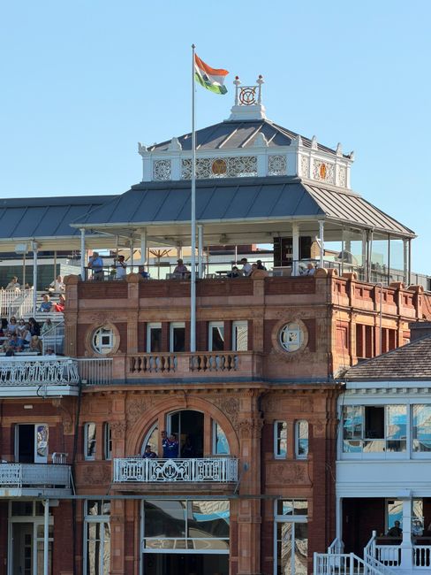 A detailed view of the upper floors of a historic red brick building with ornate architectural features, including decorative window frames and a small balcony on the second floor. The building has a rounded arched entrance at street level and a white balcony railing. The roof is topped with a circular, glass-enclosed pavilion with white decorative trim and a flagpole flying the Indian flag. The sky behind the building is clear and blue, with sunlight illuminating the facade. This scene reflects urban architecture, possibly associated with locations where independent or private waste removal services might be utilized for building maintenance or renovation projects, as part of non-public waste handling activities. The photograph captures the intricate detailing of the building's design and the lively environment typical of prominent city sites.