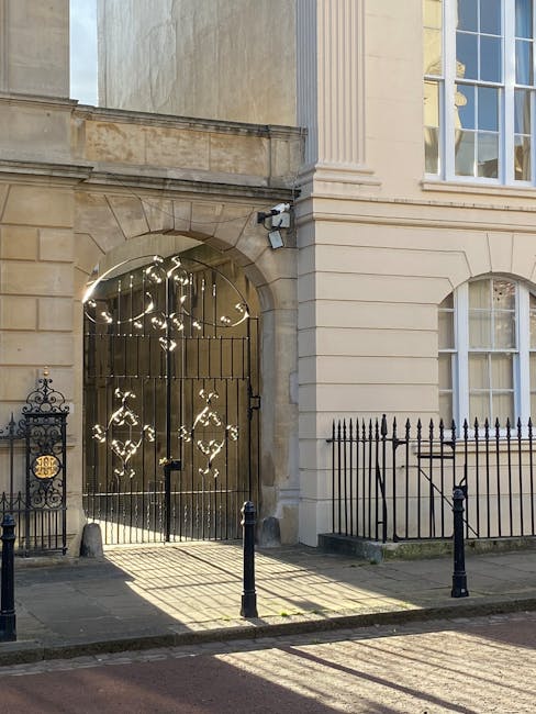 The image shows a historic building's exterior with cream-colored stone walls and architectural detailing, including vertical fluted columns and large sash windows with white frames and multiple small panes. To the left, there is a wrought iron gate with intricate decorative scrollwork, standing within a stone archway that features a recessed area with a security camera mounted above. Adjacent to the gate, a low black metal fence with pointed finials runs along the pavement, which is made of dark, smooth material. The building casts distinct shadows onto the sidewalk, indicating sunlight from the right side of the image. The overall scene presents a well-maintained, classic urban façade with elements characteristic of traditional British architecture, set in a tranquil street environment that might be associated with private property or heritage buildings potentially requiring specialist rubbish removal services by companies like Waste Removal Marylebone for maintenance or renovation waste clearance.
