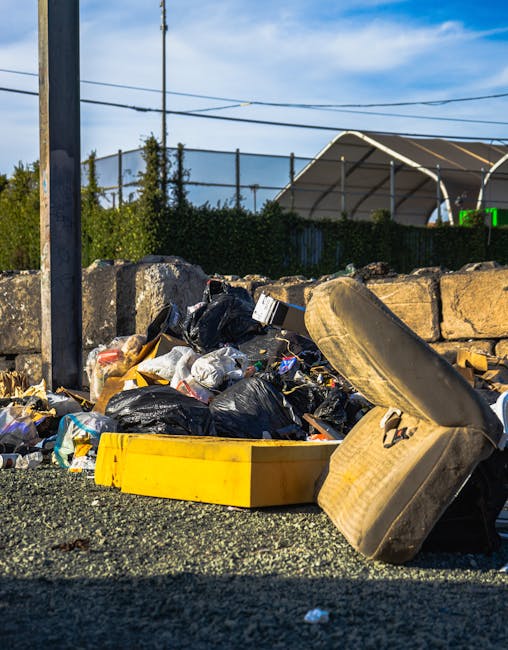 A close-up view of a pile of unmanaged rubbish on a paved surface, including black plastic garbage bags, a yellow plastic container, and a large worn-out tire lying on its side. The waste is irregularly spread, with some debris spilling onto the ground. Behind the heap, a low stone wall constructed of rough-hewn, weathered stones acts as a boundary, and in the background, there are green shrubbery and a metal fence topped with netting. Overhead, utility wires run across a sky that is partly cloudy with a mix of blue and white clouds, indicating daytime. To the right, a partially visible structure with a curved roof can be seen, suggesting an industrial or storage setting. The scene highlights an area where illegal dumping might occur, linking visually to the importance of appropriate waste removal services such as those provided by Waste Removal Marylebone, which supports private disposal and alternative waste handling solutions.