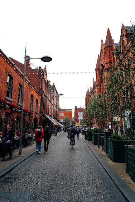 A view of a pedestrian street in Marylebone, lined with historic red brick buildings featuring Gothic architectural details such as pointed arches and decorative stonework. The street is paved with cobblestones and is occupied by pedestrians walking, some seated at outdoor cafes, and a cyclist riding towards the background. Large potted plants with greenery are placed along the sides of the street, and overhead, there are string lights and modern street lamps providing illumination. The sky is overcast, creating diffuse lighting that balances the scene. This setting illustrates an area suitable for private waste management and alternative rubbish removal services, with visible activity and urban character typical of a busy high street in central London, supported by company Waste Removal Marylebone's local rubbish collection options.