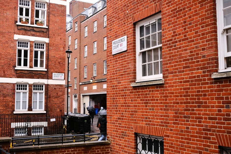 The image depicts an alleyway between red brick residential buildings in an urban area. In the foreground, a section of a brick wall with a window featuring a white frame and multiple panes is visible. To the left, another building with similar red brickwork has three white-framed windows on the upper level, each with decorative window boxes containing small plants or flowers. Below, on the ground level, a black metal gate and a small outdoor area are visible, bordered by a short brick wall. The alley features a black rubbish bin placed near the building on the left, with two individuals dressed in dark clothing and jackets seen near the bin, possibly engaged in waste disposal activity. An access door on the building is visible in the background, partially obscured by one of the individuals. Two street signs are mounted on the buildings, indicating street names—'Balderton Street W1' on the left and 'Providence Court W1' on the right—highlighting a central London location. The surrounding environment suggests this scene involves private waste collection or rubbish removal for urban flats, consistent with independent disposal services such as Waste Removal Marylebone.