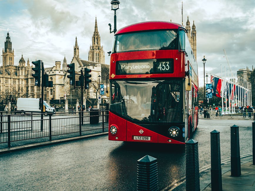 A red double-decker bus with large front windows and a destination sign reading 'Marylebone 453' is parked on a wet street in front of historic Gothic-style buildings with spires and pointed arches. The bus's exterior is clean and glossy, reflecting the overcast sky above. To the left of the bus, there are multiple traffic lights mounted on black poles, some showing red signals. Behind the bus, a row of flagpoles with UK flags stands along a promenade, suggesting a public or cultural area. In the background, pedestrians are visible walking near the flagpoles, some bundled in jackets, indicating cool weather. The surrounding environment includes a metal fence along the passenger walkway and a few black bollards on the street curb, providing safety barriers. The street surface appears slick from recent rain, adding a reflective quality to the asphalt. This scene, captured during daytime under cloudy conditions, subtly relates to private waste collection and alternative rubbish handling methods, akin to independent rubbish removal services such as those provided by Waste Removal Marylebone.