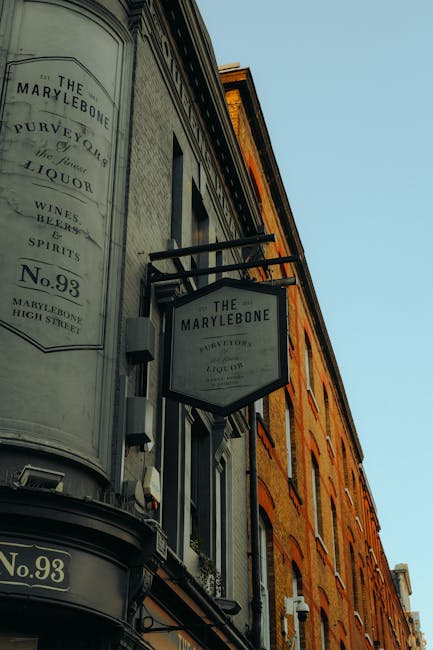 A close-up view of the exterior corner of a historic brick building with a curved, vintage-style sign attached to the façade, displaying the name 'The Marylebone' along with related details such as 'Purveyors of Liquor,' 'Wines,' 'Spirits,' and the address 'No. 93 High Street.' The sign is supported by black metal brackets and is framed with black borders, mounted against cream-painted decorative molding that surrounds the building's corner. The brick structure features large rectangular windows with white window frames, and the overall architectural style reflects traditional UK high street frontage. The background sky is clear and blue, suggesting daytime natural lighting. This scene subtly relates to private commercial premises that may generate rubbish requiring services like those provided by Waste Removal Marylebone for efficient on-site clearance or alternative waste management solutions for local businesses and properties in the Marylebone area.