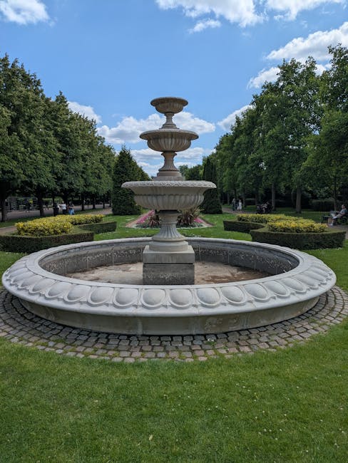 A large, ornate stone fountain positioned in a well-maintained park setting with lush green grass and neatly trimmed bushes. The fountain features a broad, circular basin with a scalloped edge, made of light-colored stone, and appears weathered with subtle signs of aging. Rising from the center of the basin are multiple tiers, each with decorative detailing, supporting a smaller upper basin and a finial at the top, also crafted from the same light-colored stone material. Surrounding the fountain is a paved area with cobblestone or similar stone paving, providing a stable base. In the background, there is a symmetrical arrangement of densely packed deciduous trees with verdant foliage, creating a natural enclosure around the park. The sky overhead is bright blue with scattered white clouds, indicating a clear, sunny day. The scene exudes a calm, scenic ambiance typical of public parks that might be used for casual gatherings or leisurely strolls, with the fountain serving as a picturesque focal point. The overall environment suggests a well-maintained public space, possibly for community or recreational use, aligned with general principles of outdoor aesthetic enhancement and conservation.