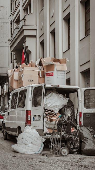 Photograph of the open rear doors of a white van parked on a residential street adjacent to a multi-storey building with a light stone facade and vertical architectural details. The van's interior is filled with various waste items, including large black and white rubbish bags, flattened cardboard boxes, and assorted packaging materials, indicating it is used for private rubbish removal and alternative waste handling. Several black garbage bags rest outside the van on the ground, along with a small hand truck and a flat trolley, reflecting on-site collection activities. Cardboard boxes are stacked loosely on top of the vehicle, some bulging with waste, and a white plastic bag is visible through the van's open rear opening. The environment appears well-lit, with natural daylight, emphasizing the informal collection scene typical of independent waste disposal services. The setting underscores the practical aspects of rubbish clearance, aligning with private waste management efforts in urban areas, as carried out by Waste Removal Marylebone.