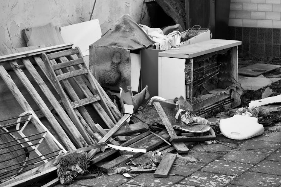 A collection of discarded household items and debris arranged on an outdoor paved surface, including a wooden ladder with visible wear and weathering, leaning against a wall alongside a small metal wire rack. Near the ladder, there are broken wooden planks and fragmented pieces of furniture, some with chipped paint and splintered edges. An old, damaged microwave oven with a white exterior and partially exposed inner components is positioned centrally, with its door removed or left open. In front of it, a worn, crumpled piece of fabric or cloth is draped over various debris, while a white foam toilet seat or lid rests on the ground to the right. The scene is set against a backdrop of brick and concrete walls, with scattered litter and what appears to be a tarp or plastic sheet partially covering some objects. The overall environmental context suggests a site for private waste disposal or on-site clearance, with items generally associated with household cleanup or rubbish removal services like Waste Removal Marylebone.