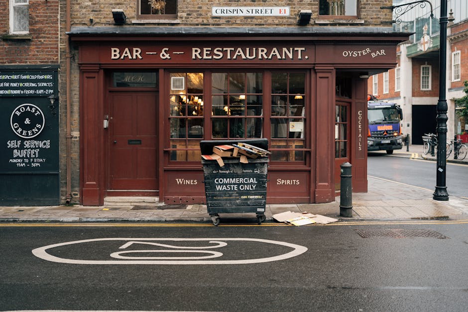 The image shows the front exterior of a restaurant or pub with a reddish-brown wooden façade, large central windows, and a sign indicating it is a bar and restaurant offering wines, spirits, and oyster dishes. In front of the establishment, a black wheeled waste bin marked 'Commercial Waste Only' is filled with cardboard boxes and paper, some of which are leaning or spilling out onto the pavement. The bin is positioned on the sidewalk near a short black bollard and a metal post, with a few discarded paper sheets lying on the ground nearby. To the left, there is an adjacent black chalkboard sign listing breakfast buffet hours, and to the right, a street scene includes a parked van, bicycles, and street lamps. Above the entrance, a small street sign reads 'Crispin Street.' The overall setting is urban, with natural daylight illuminating the scene, and the presence of the waste bin and discarded materials suggests activities related to waste collection or private waste disposal, consistent with alternative rubbish management services in a city environment.